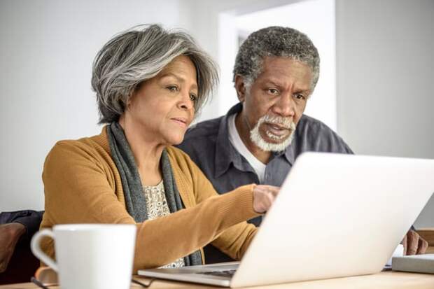 A couple reading content on a shared laptop while seated at a table in their home.