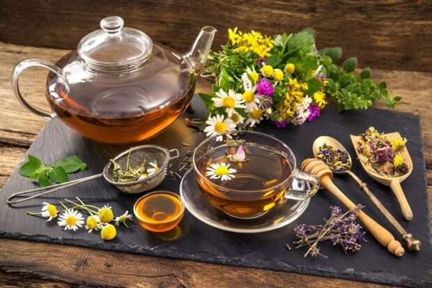 Стеклянный чайник, прозрачная чашка чая с плавающим цветком ромашки среди сухих и свежих трав, на переднем плане ситечко, пиала с мёдом, ложка, лопаточка для мёда и трав