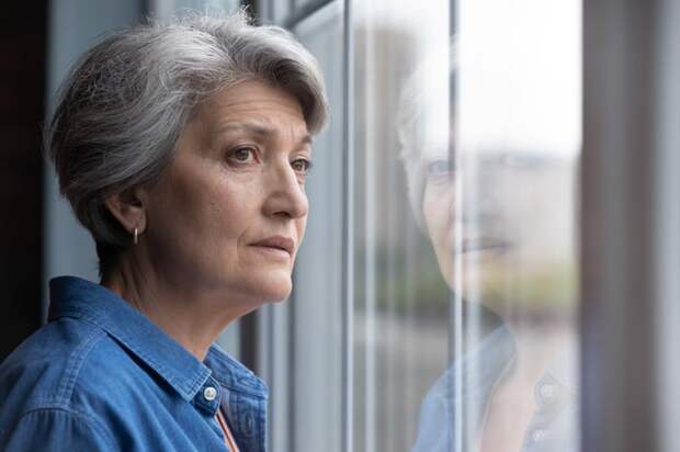 An older person gazes out of a window with their expression filled with worry.