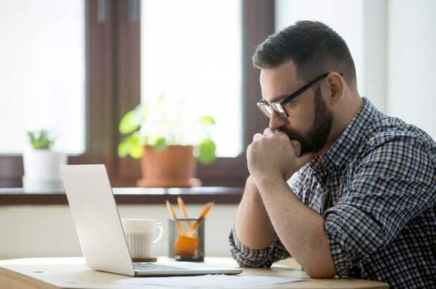 A man sitting at a desk with his laptop open and looking stressed with his chin in his hands.