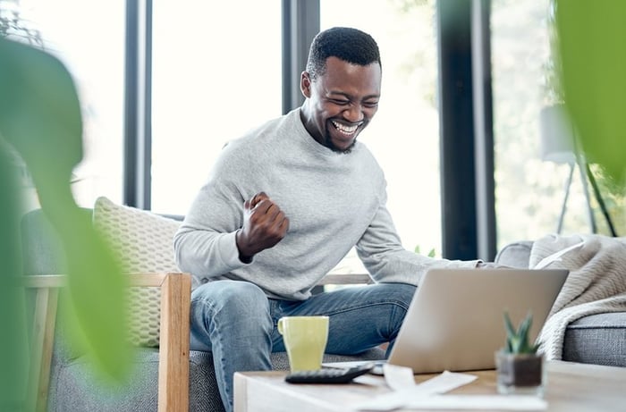 A young adult pumps their fist in celebration while going over paperwork and using a laptop at home.