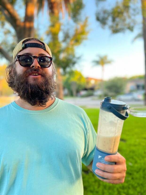 Beard man in sunglasses and a backwards hat holding a Ninja Blast Portable Blender in a city park after making a delicious peach smoothie with it.