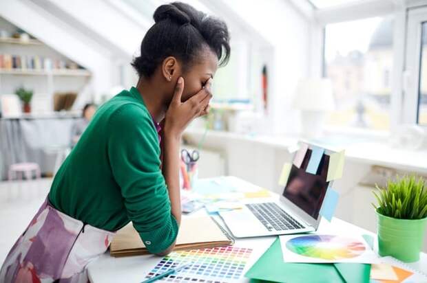 A stressed young woman sitting at her desk in front of a laptop with her face in her hands.