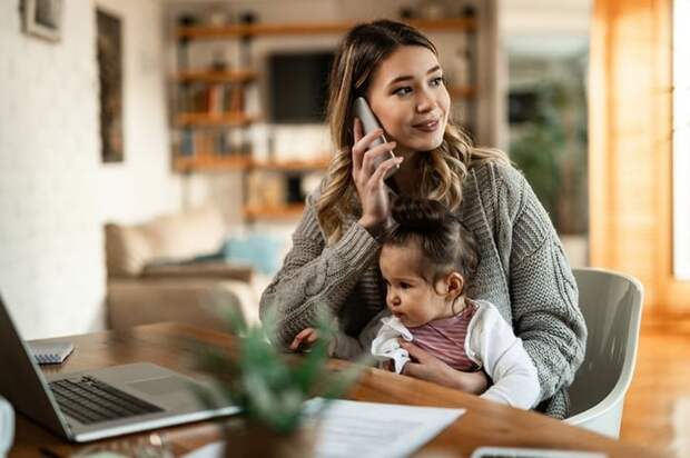 A woman sitting at a desk at home with her baby in her lap while making a phone call.