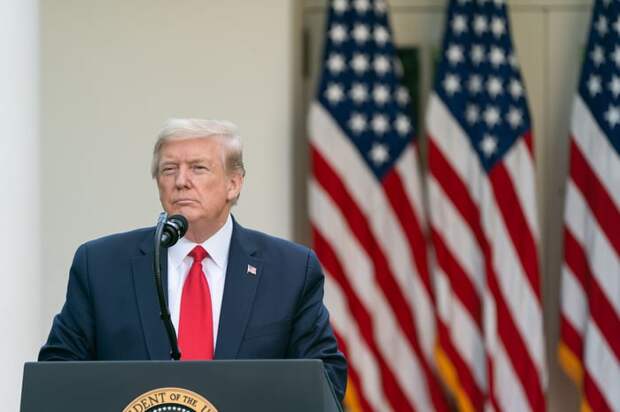 Donald Trump stands at a podium listening to a report with American flags in the background.