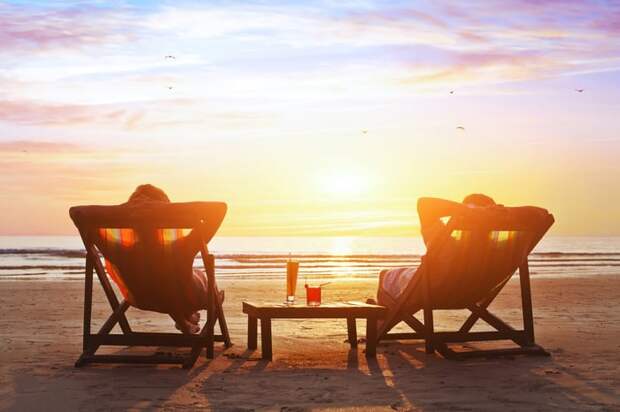 Two people sitting on lounge chairs on a beach looking at the sunset.