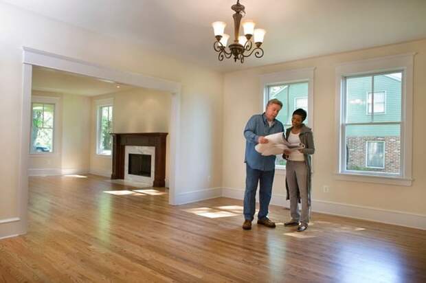 A man and woman standing in an empty living room and looking at blueprints of the house.