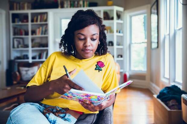 A person sitting in a living room chair and writing in a colorful notebook.